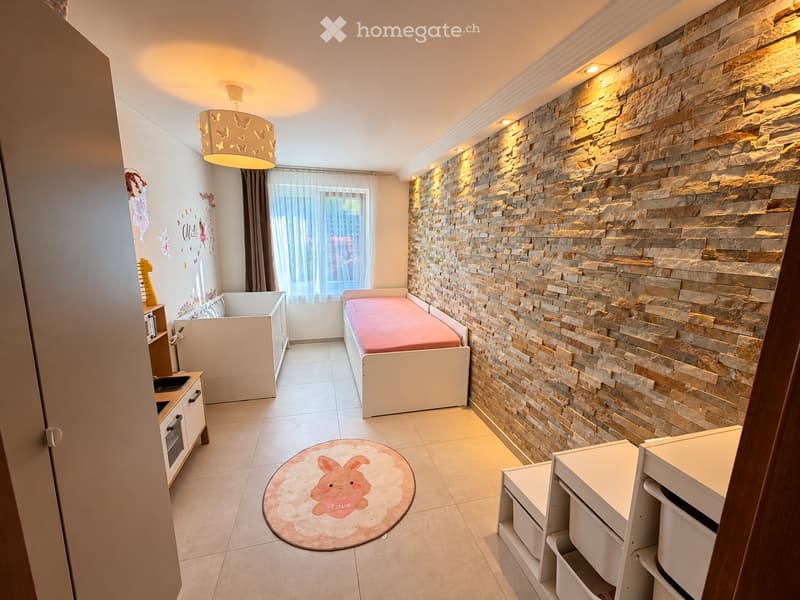 Cozy children's bedroom with a stone accent wall, a single bed with a red blanket, a small cabinet, a round rug with a bunny design, and a storage unit.