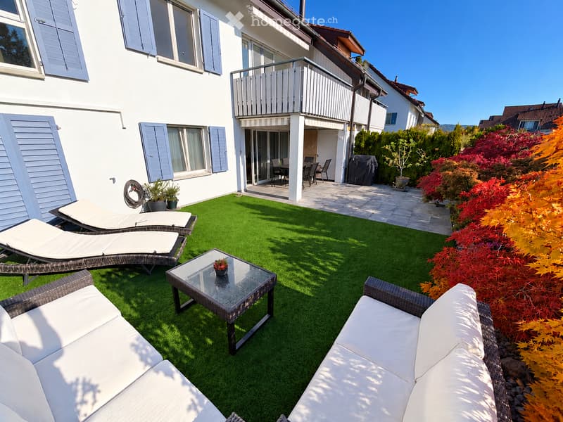 Well-maintained garden with artificial grass, white sofa, glass coffee table, sun loungers, and colorful foliage.