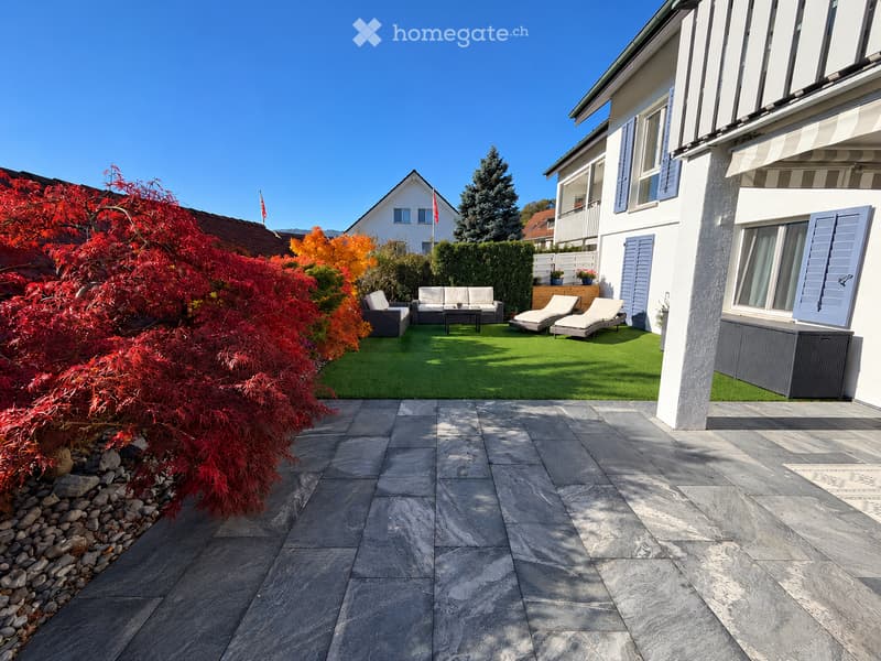 Well-maintained garden with artificial grass, patio furniture including a sofa, coffee table, and chairs, surrounded by colorful foliage and a modern house.