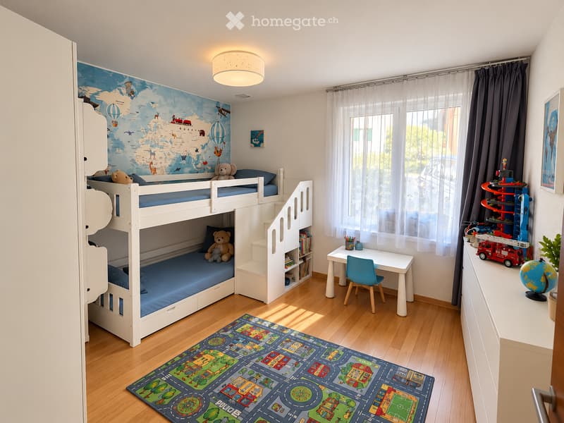 A well-lit children's bedroom with a bunk bed, a desk with a chair, a colorful play mat, and various toys including a globe, toy cars, and a toy train set. The room features a world map mural on one wall and large windows with sheer curtains.