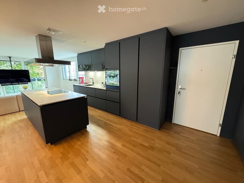 Modern kitchen with dark grey cabinets, a central island, and wooden flooring. Features include a stand mixer, range hood, and large windows with a view of greenery.