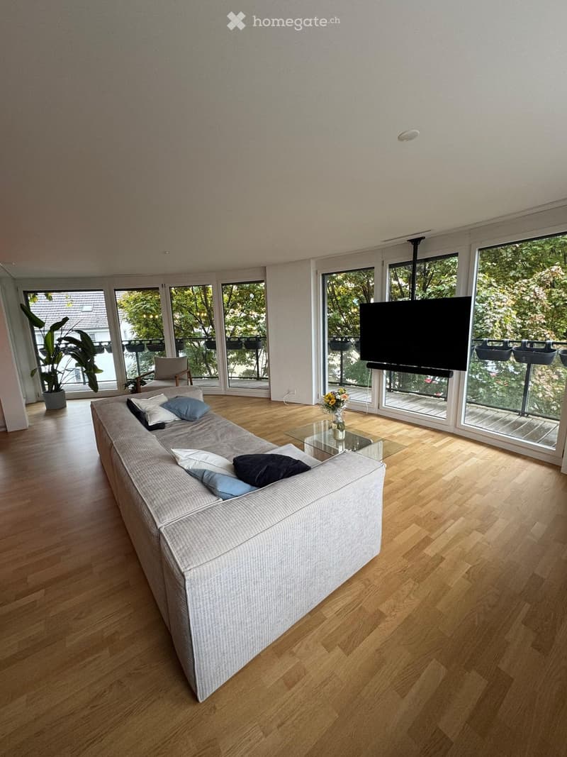Modern living room with light wooden flooring, large windows, and a balcony. Features a grey sofa with pillows, a glass coffee table, a vase with flowers, a chair, and a television mounted on the wall.