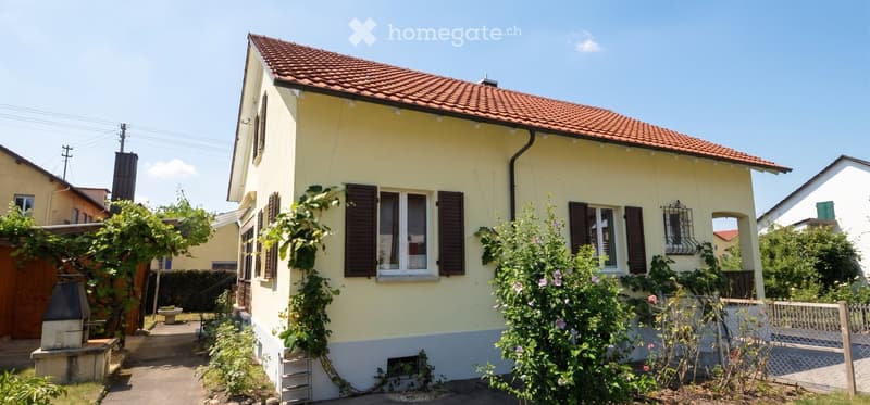 Two-story yellow house with red-tiled roof, multiple windows with shutters, and surrounding greenery.