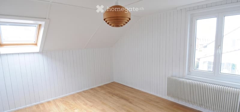 Empty attic room with white paneled walls, wooden floor, skylight window, large windows, and a hanging wooden pendant light.