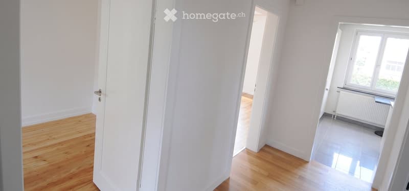 Empty hallway with white walls, wooden flooring, and a window with radiator. No furniture or equipment present.