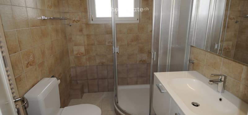 Bathroom with beige tiled walls and floor, white toilet, sink with chrome faucet, and a glass-enclosed shower area.