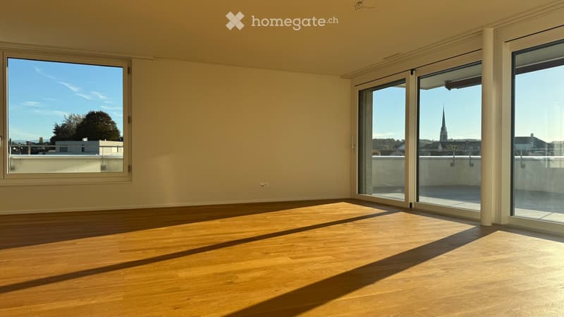 Empty room with wooden flooring and large windows, offering a view of a cityscape and a church spire.