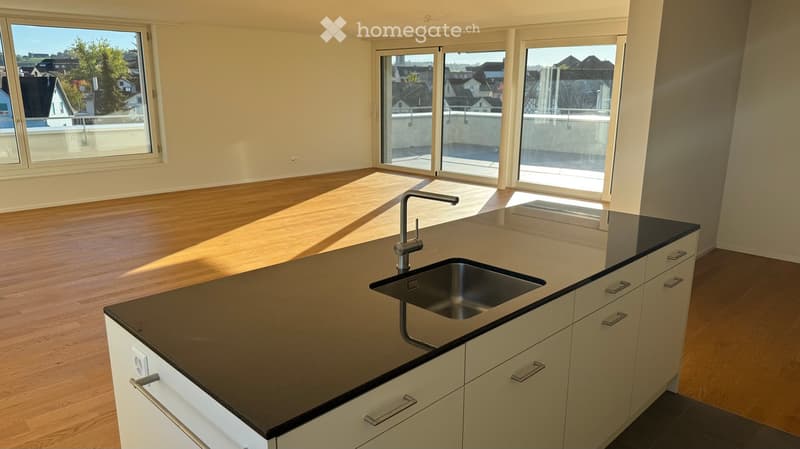 Empty kitchen with wooden flooring, white cabinets, black countertop, and a stainless steel sink. Large windows and sliding doors lead to a balcony.