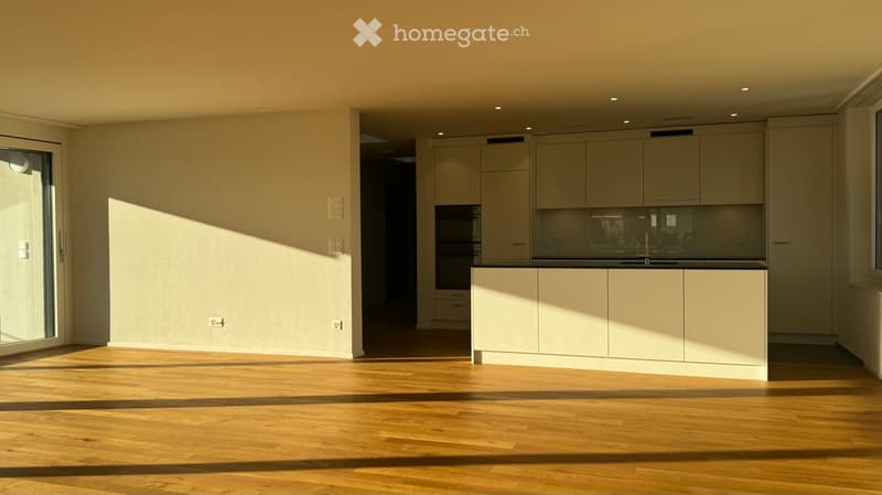 Empty room with light wooden flooring, white walls, and built-in kitchen cabinets visible in the background. Sunlight streams in from a large window on the left side.