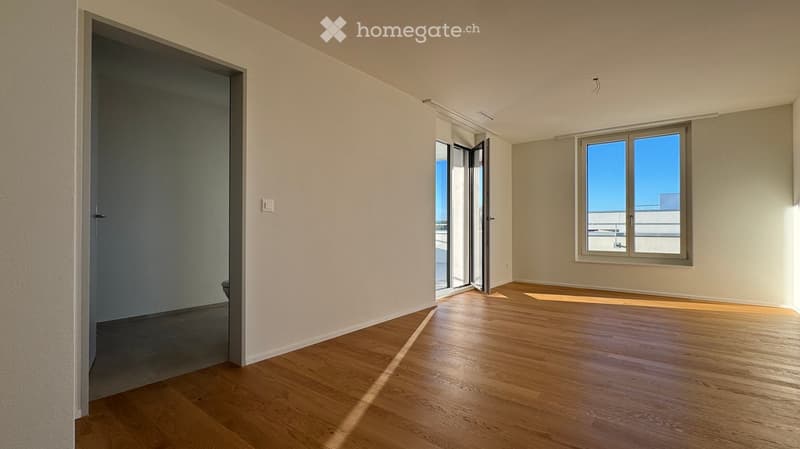 Empty room with wooden flooring, white walls, and large windows with a view of the outside. The room features a sliding glass door leading to a balcony area.