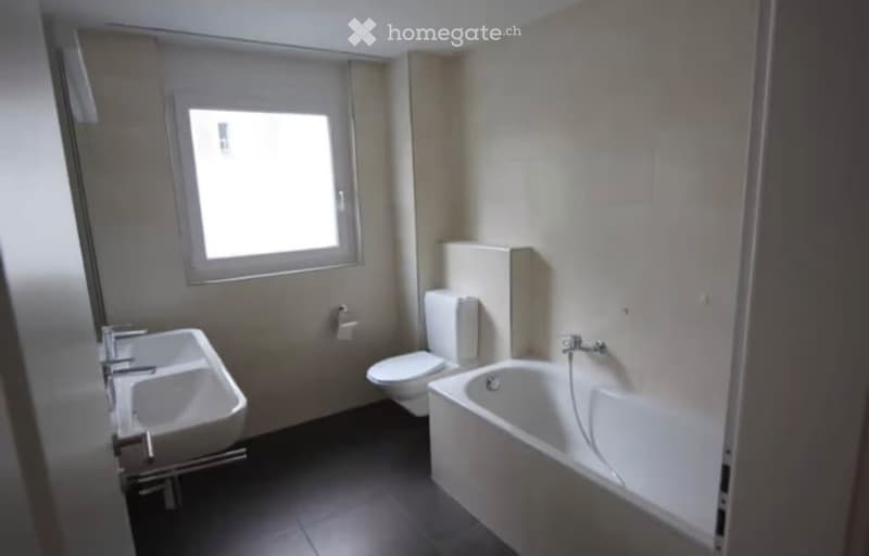 Modern bathroom with a white sink, toilet, and bathtub, featuring a window for natural light and a dark tiled floor.