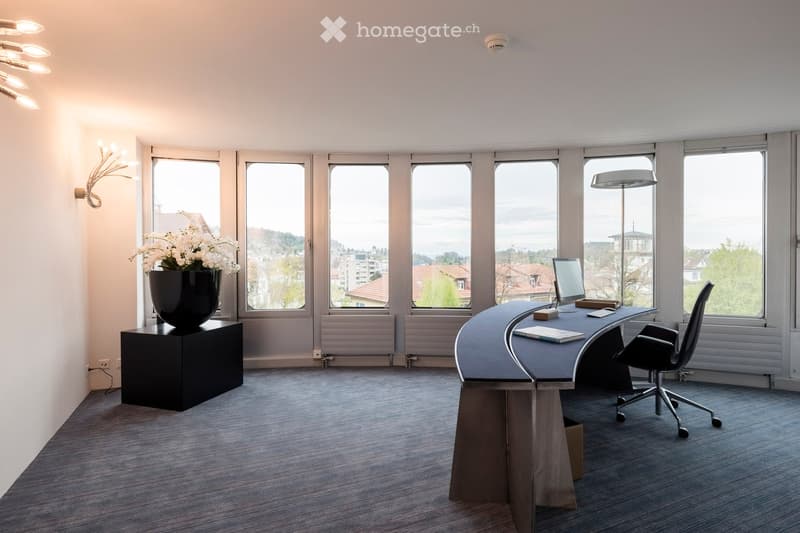 Modern office space with large windows offering a city view, featuring a sleek curved desk, ergonomic chair, computer monitor, vase with white flowers, and wall-mounted and floor lamps.