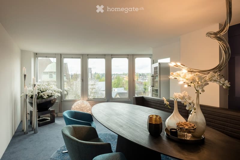 Modern living room with large windows offering a city view, featuring a wooden table, two teal chairs, decorative vases, a candle holder, and a flower arrangement.