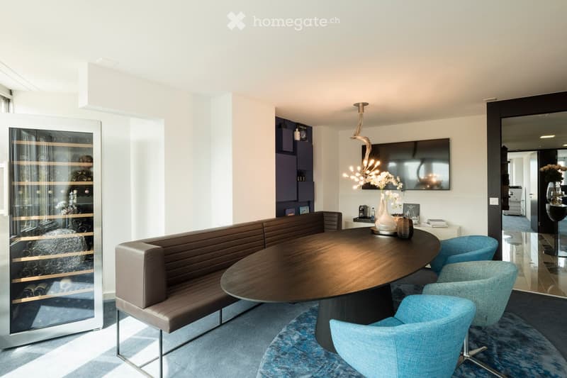 Modern living room with a round wooden table, leather sofa, blue chairs, wine cabinet, decorative vase, and a television mounted on the wall.