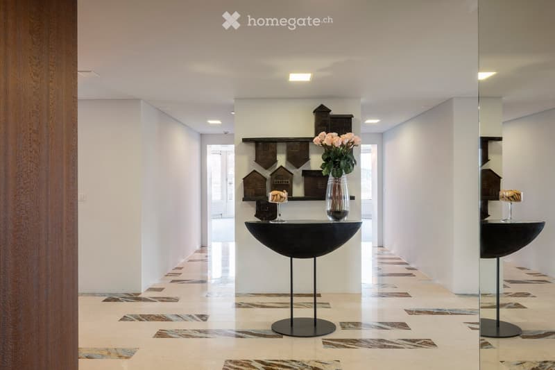 Modern hallway with white walls, patterned tile floor, recessed lighting, and decorative shelves with small house models and a vase of flowers on a black console table.