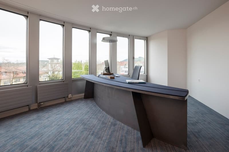 Modern office space with large windows, a sleek black desk, a chair, a computer, a lamp, and a book on the desk.