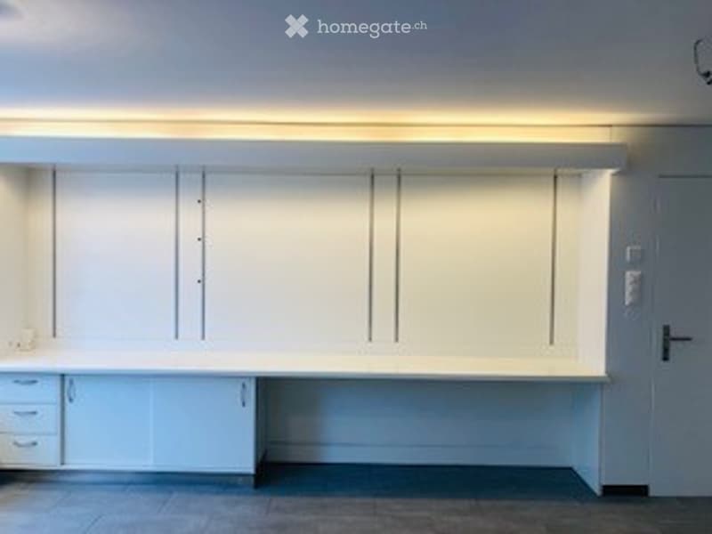 A clean, modern unfurnished room with white cabinetry, a long countertop, and built-in storage. The space features sleek, minimalist design with a neutral color palette and bright lighting.