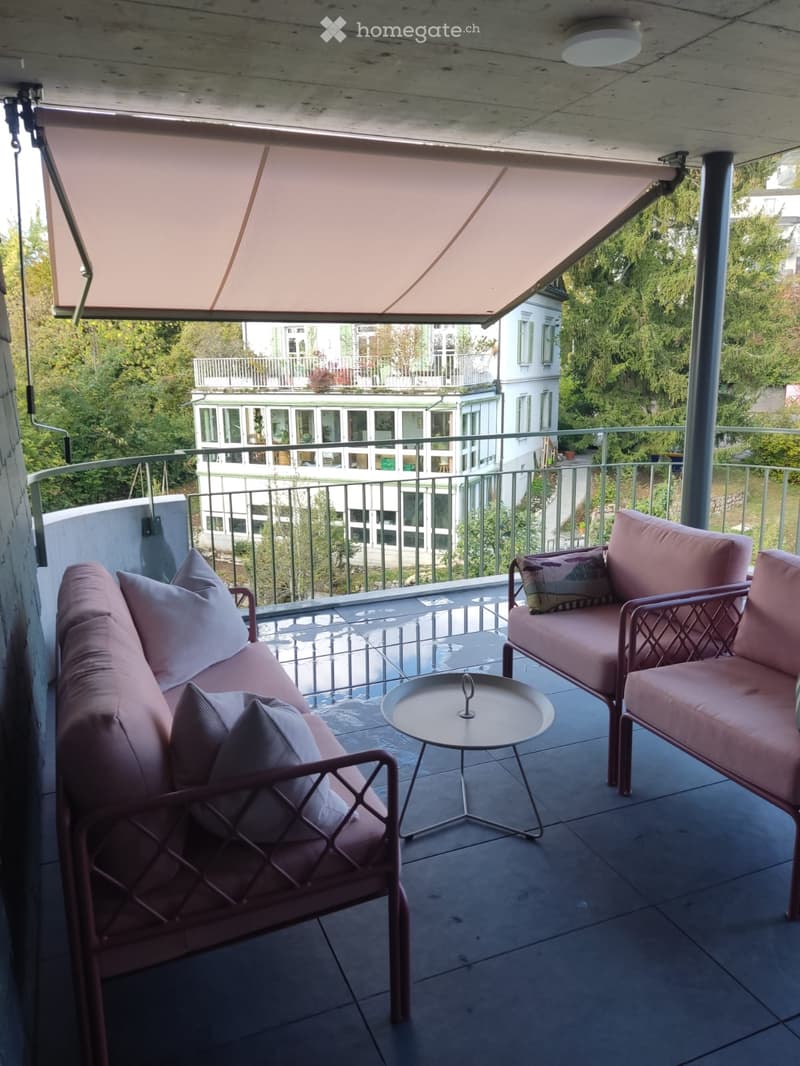 Balcony with pink sofas, chairs, and a small round table, featuring a retractable awning and a view of neighboring buildings and greenery.