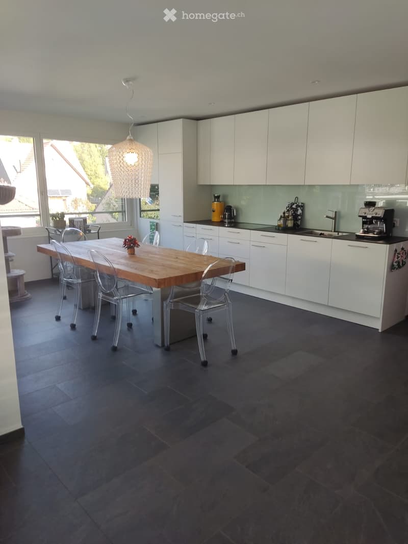 Modern kitchen with white cabinets, dark tiled floor, wooden dining table with transparent chairs, and various appliances including a coffee maker and sink.