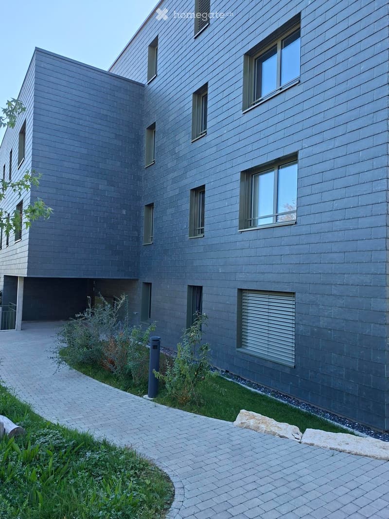 Modern two-story house with grey wooden siding, multiple windows, and a small garden area with a paved walkway.