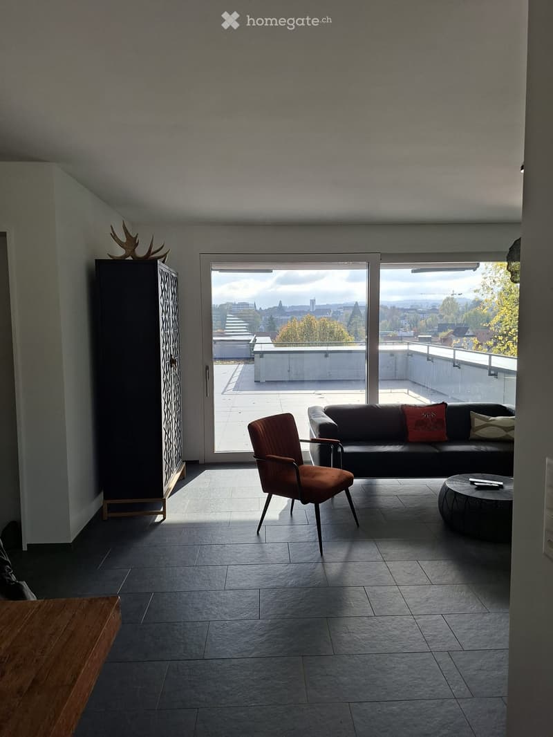 Modern living room with large windows offering a scenic view, featuring a dark grey sofa with colorful cushions, a round coffee table, and a wooden side table.