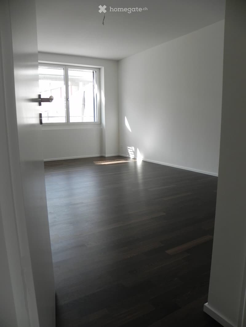 Empty room with wooden flooring and white walls, featuring a large window with blinds allowing natural light to enter.