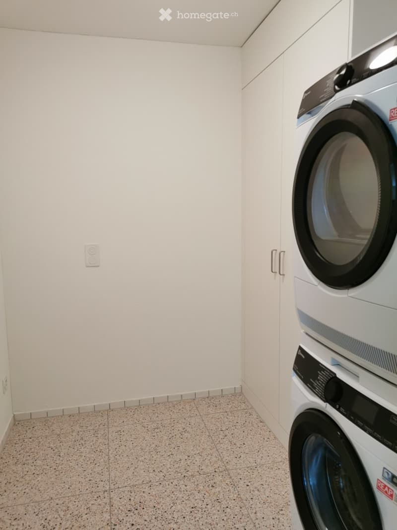 A small, clean utility room with a washing machine and dryer stacked vertically, white walls, and a tiled floor.