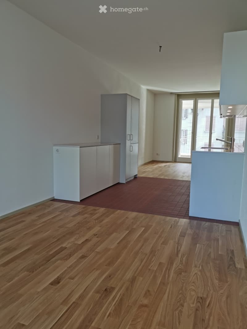 Empty kitchen with wooden flooring, white cabinets, and a sliding glass door leading to a balcony.