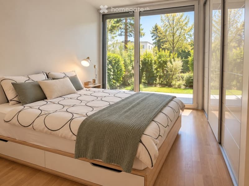 Modern bedroom with a large bed, multiple pillows, a green throw blanket, a nightstand, and a wall-mounted lamp. Large windows offer a view of greenery outside.