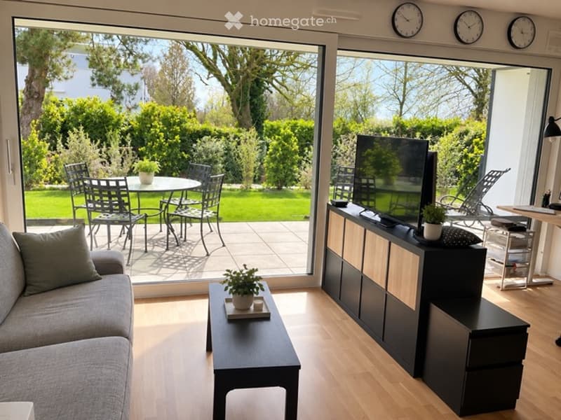 Bright living room with large windows overlooking a garden, featuring a grey sofa, black coffee table, TV stand, and outdoor dining set.