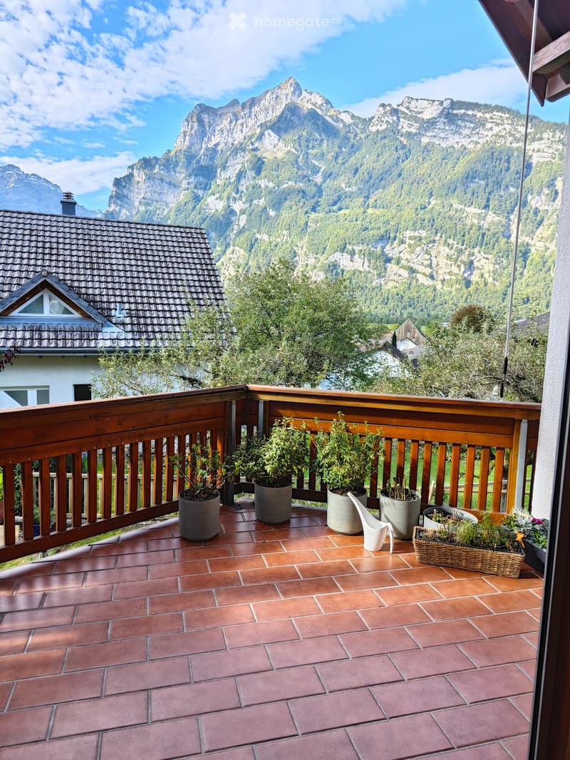 Balcony with wooden railing, terracotta tiles, and potted plants, overlooking a mountain landscape.