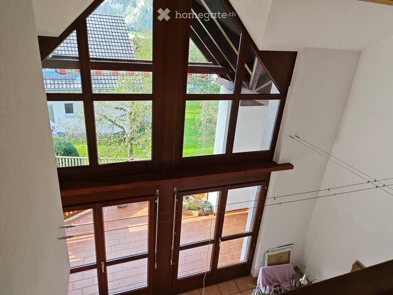 Bright, airy room with large windows offering a scenic view of the surrounding landscape. The room features a wooden door leading to a tiled patio area, a small table with a plant, and a chair with a pink cover.