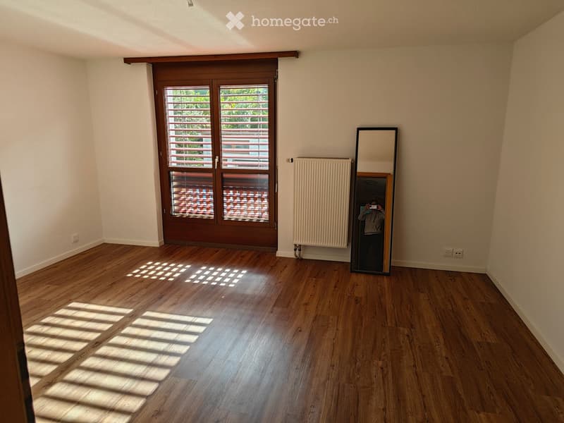 Empty room with wooden flooring, large window with blinds, radiator, and a mirror on the wall.