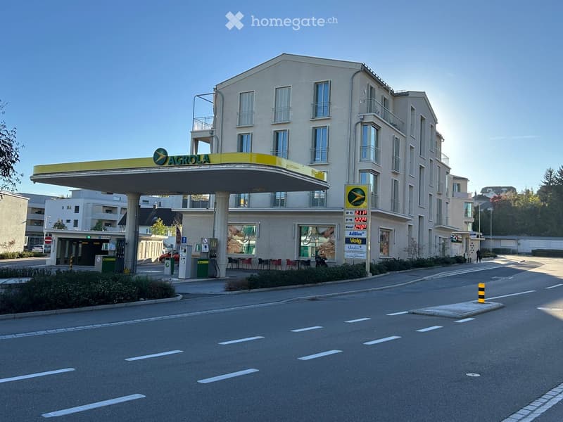Multi-story building with a modern facade, featuring balconies and large windows. A gas station with a prominent 'AGROLA' sign is attached to the building.
