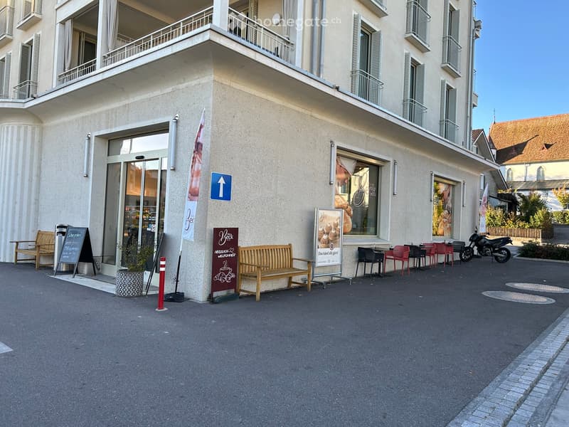 Facade of a multi-story building with balconies, a storefront, outdoor seating area, and a motorcycle parked nearby.