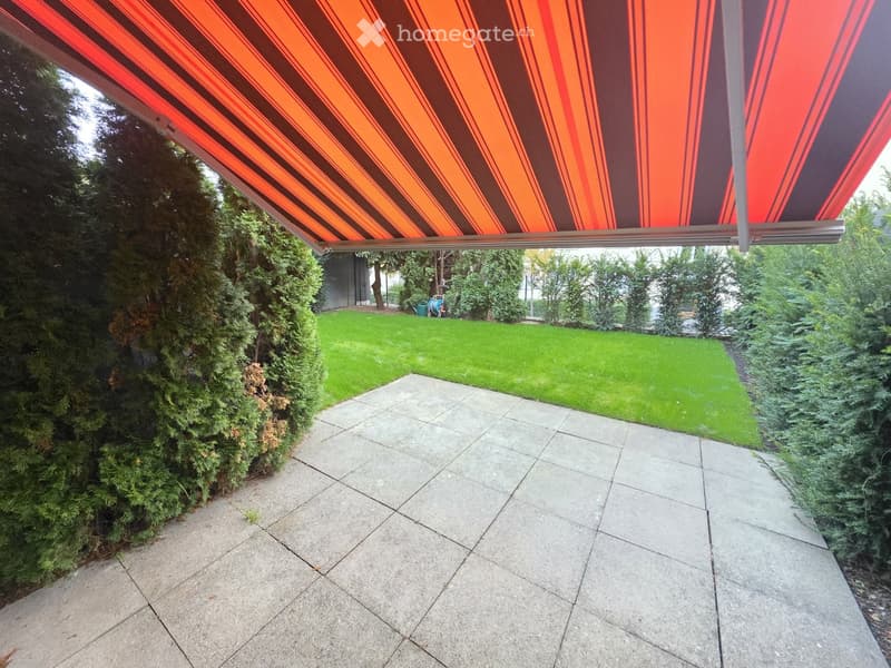 A paved patio area with a large retractable awning, surrounded by green bushes and a well-maintained lawn.