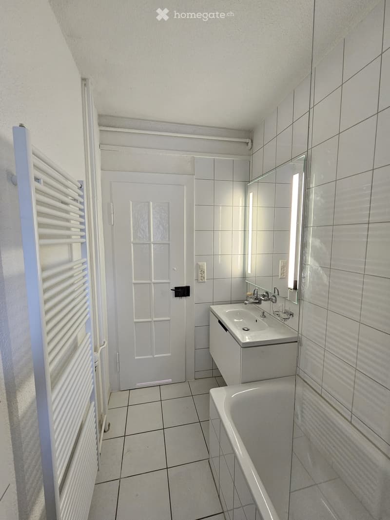 Modern bathroom with white tiled walls and floor, featuring a sink, bathtub, mirror, and radiator.