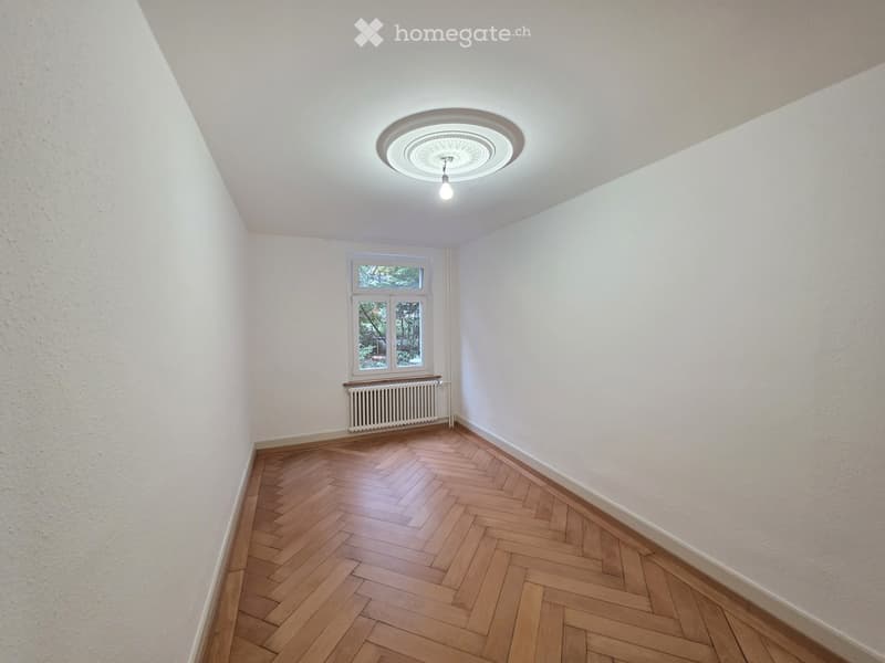 Empty room with wooden herringbone flooring, white walls, a single window with a radiator below, and a ceiling light fixture.