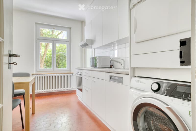 Bright kitchen with white cabinets, a sink, microwave, dishwasher, and washing machine. A small dining area with a table and chairs is visible near the window.