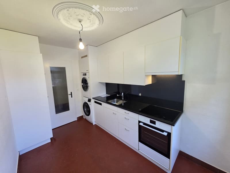 Modern kitchen with white cabinets, black countertops, stainless steel sink, oven, and washing machine. Features a hanging light fixture and a red tiled floor.