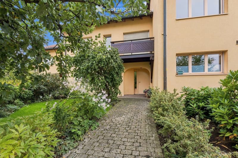 Garden path leads to a house entrance with a balcony above, surrounded by lush greenery and flowering plants.