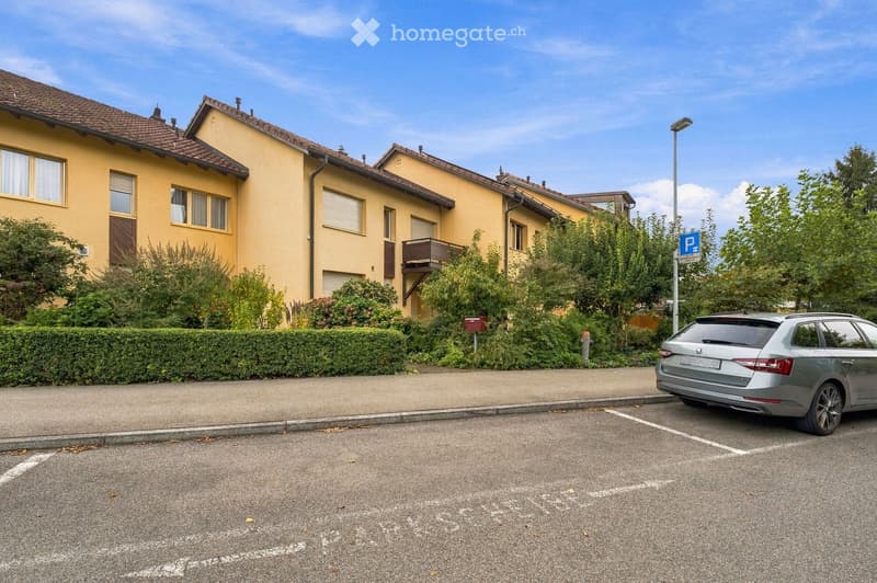 Two-story yellow house with brown roof, multiple windows, and a small balcony. A silver car is parked in a designated parking space in front of the house.