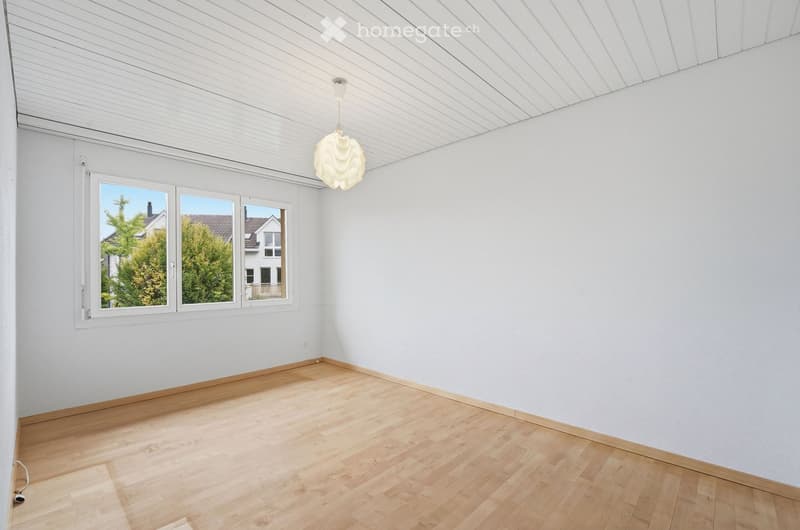 Empty room with light wooden flooring, white walls, and a large window offering a view of trees and neighboring houses.