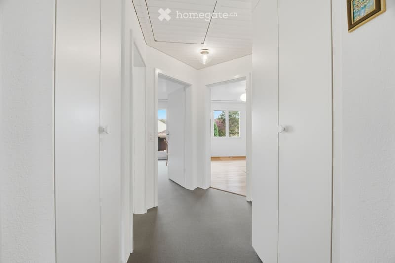 A clean, modern hallway with white walls, grey carpet, and several open doors leading to other rooms. Natural light streams in from the end of the hallway, and the ceiling features recessed lighting.