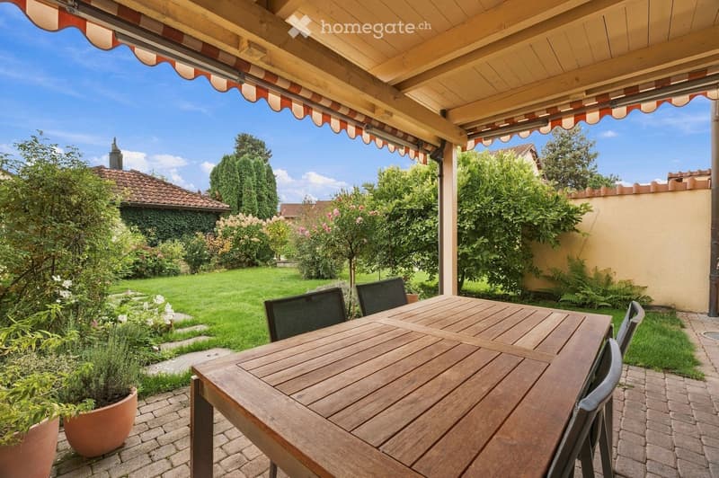 A covered outdoor patio with a wooden table and chairs, surrounded by a well-maintained garden with various plants and trees.