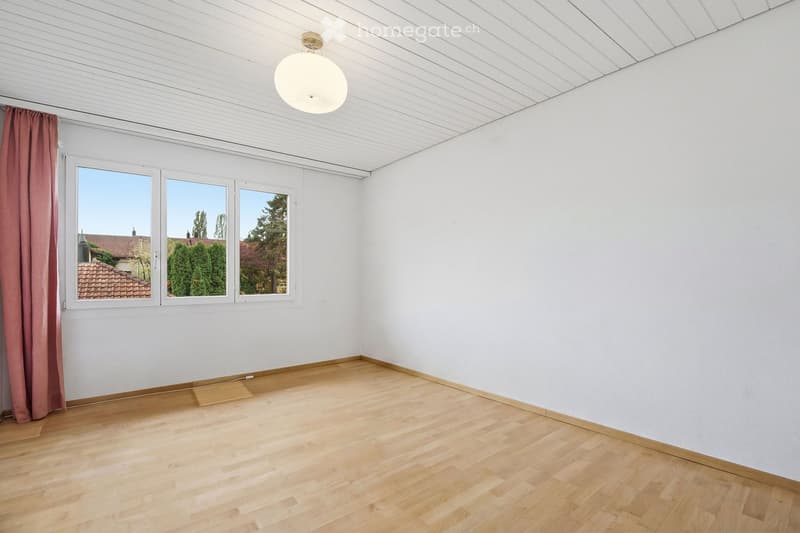 Empty room with wooden flooring, white walls, and large windows with red curtains. The ceiling has a light fixture.