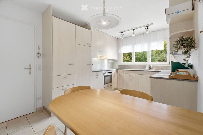 Modern kitchen with light-colored cabinets, a round wooden dining table with chairs, built-in oven, refrigerator, and sink area. Large windows provide natural light.