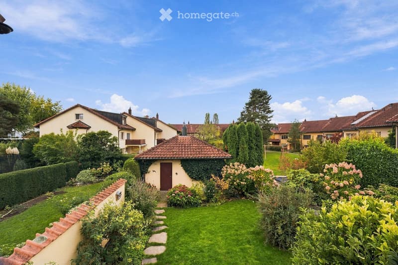 Well-maintained garden with a stone pathway, lush green grass, flowering shrubs, and a small shed surrounded by trees and neighboring houses.