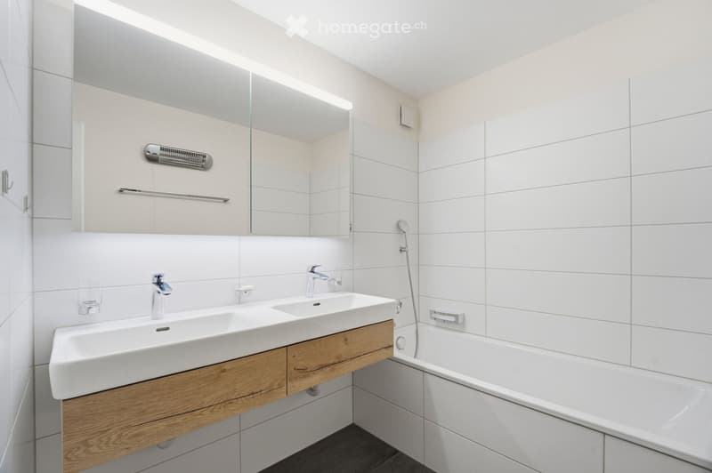 Modern bathroom with white tiles, double sink, wooden cabinet, bathtub, and large mirror with built-in lighting.