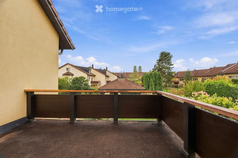 Balcony with wooden railing, view of neighboring houses and greenery, clear blue sky.